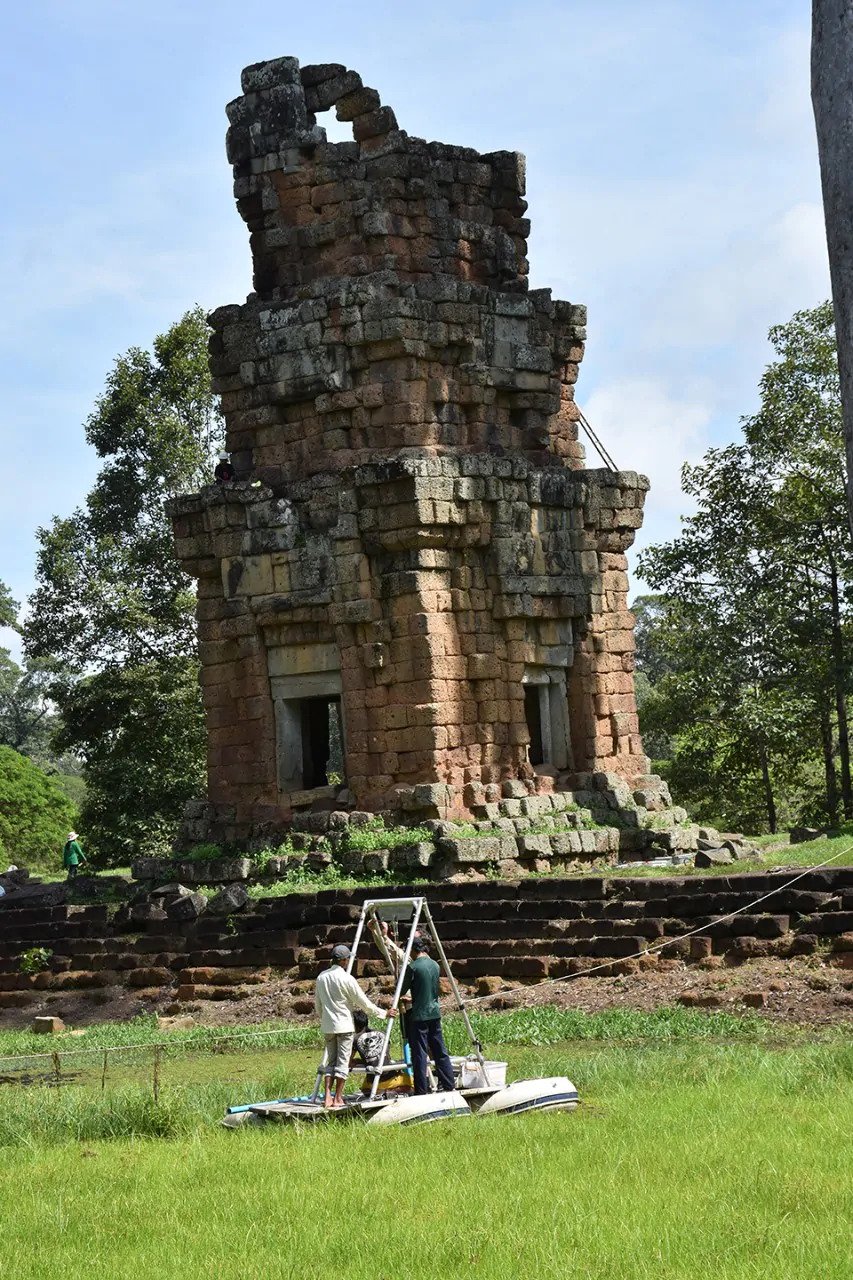 On a floating platform, Penny and team take a drill core sample from an overgrown reservoir. The 12th century tower S1, of the Prasat Suor Prat group, looms in the background.